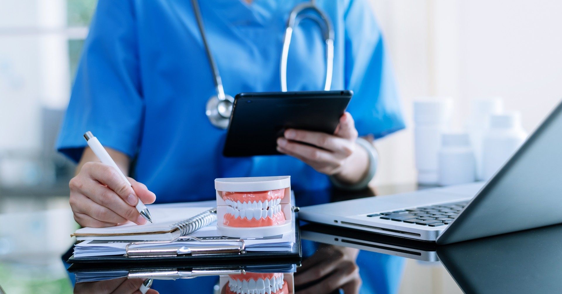 Dentist sitting at table with jaw samples tooth model and working with tablet and laptop in dental office professional dental clinic.